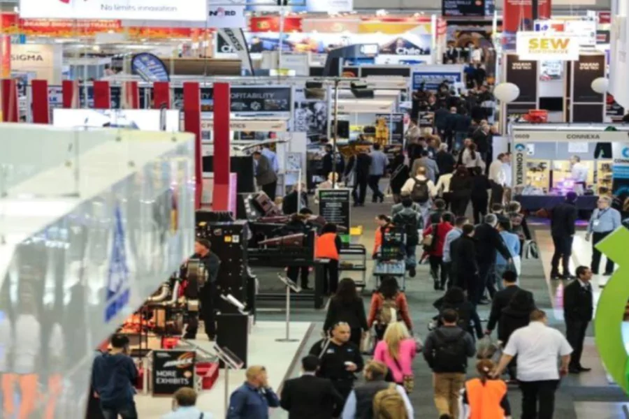 Busy scene from the AIMEX trade show, with attendees walking through rows of industrial and mining equipment exhibits under bright signage.
