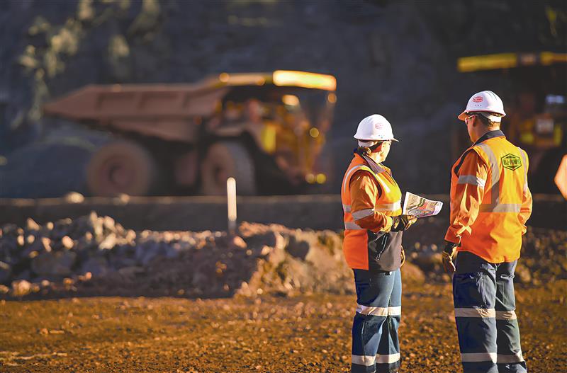 Two workers in high-vis orange jackets and white hard hats review documents at a mining site, with large dump trucks operating in the background.