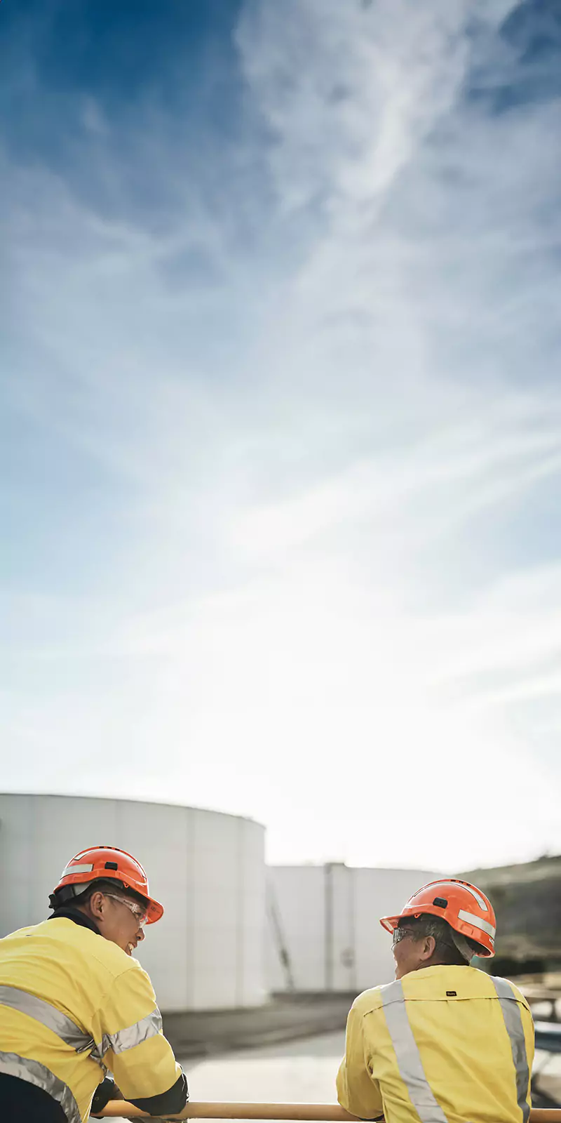 Two workers in yellow high-visibility jackets and orange helmets stand near industrial storage tanks, talking under a bright sky.