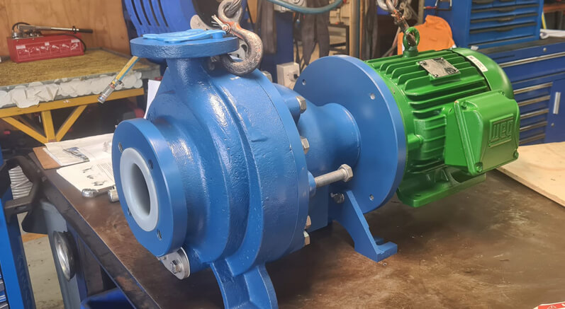Technician assembling a blue Finish Thompson Inc. (FTI) chemical transfer pump with green WEG motor on a workshop bench. The pump is designed for safe handling of acids, solvents, and corrosive liquids in chemical manufacturing plants.