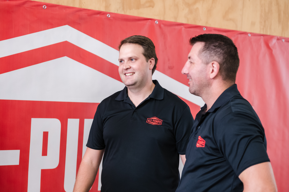 Two All-Pumps team members smiling and talking in front of a branded red company banner.
