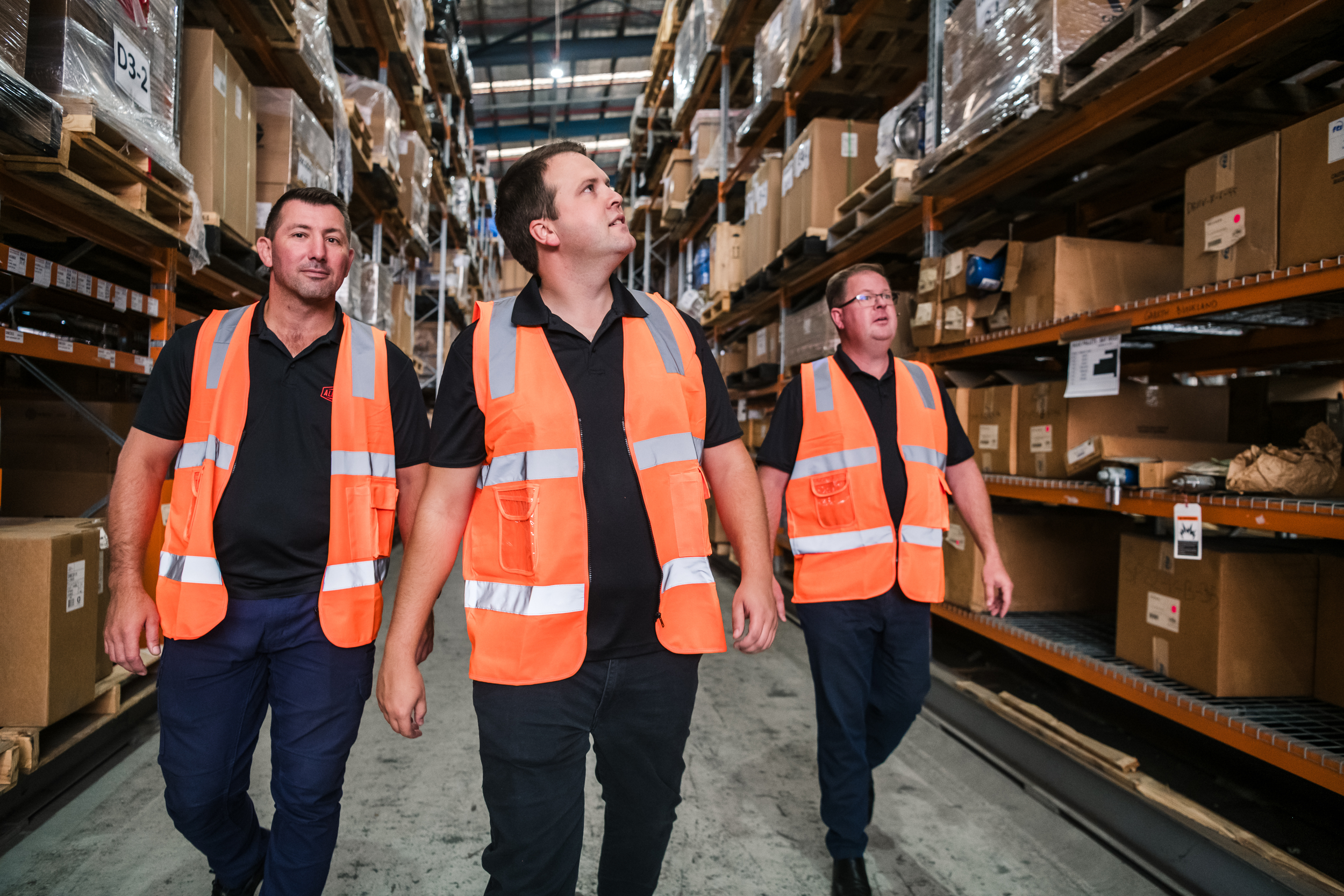 Three All-Pumps team members in high-vis vests walking through a warehouse filled with shelves of boxed inventory.