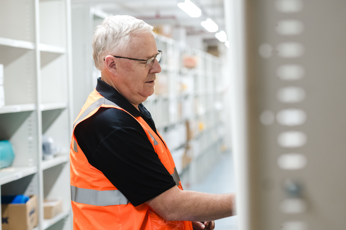 Warehouse worker in an orange safety vest inspecting items on a shelf in a well-organized storage area.