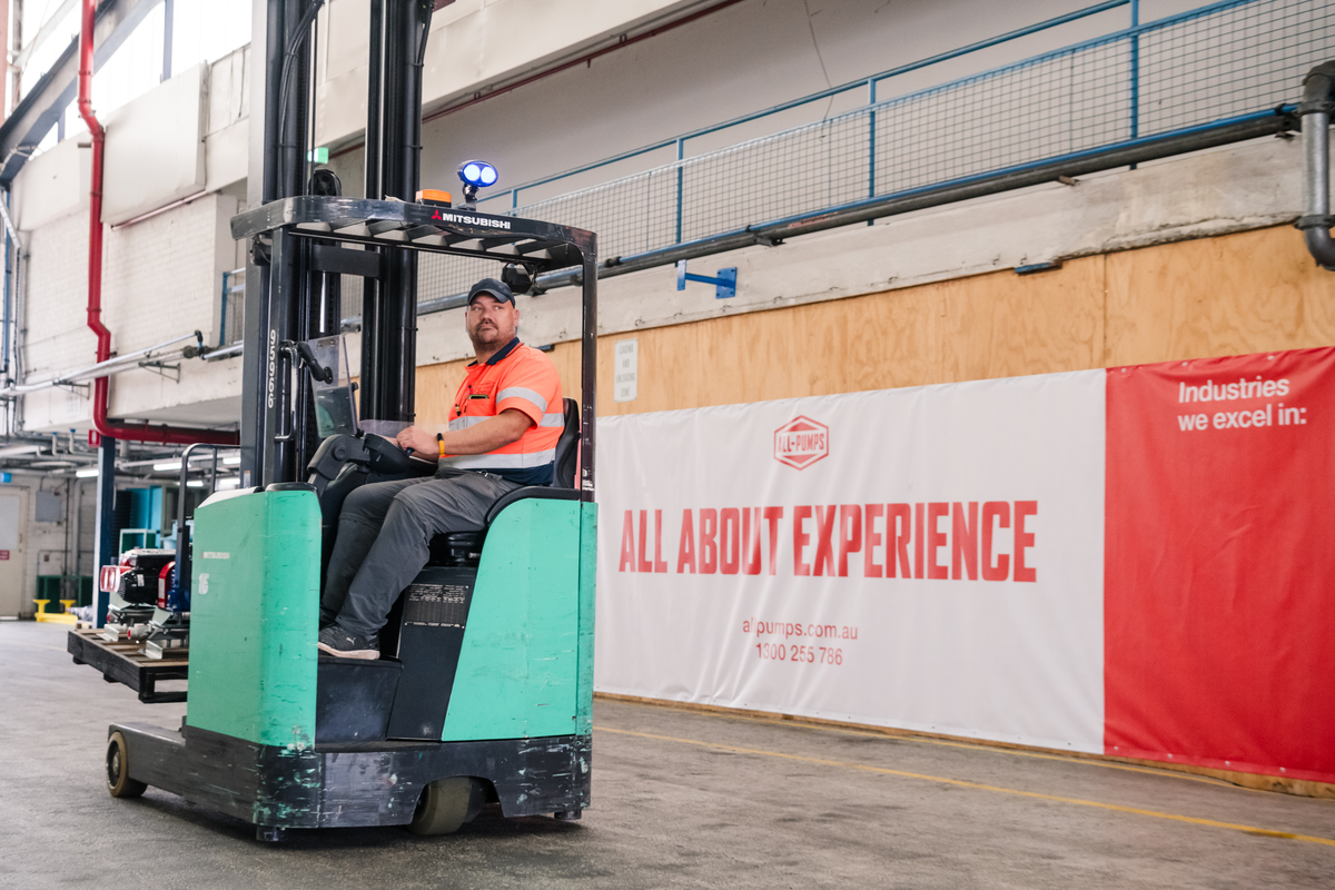 A warehouse worker drives a green Mitsubishi forklift past a large All-Pumps banner that reads "All About Experience."