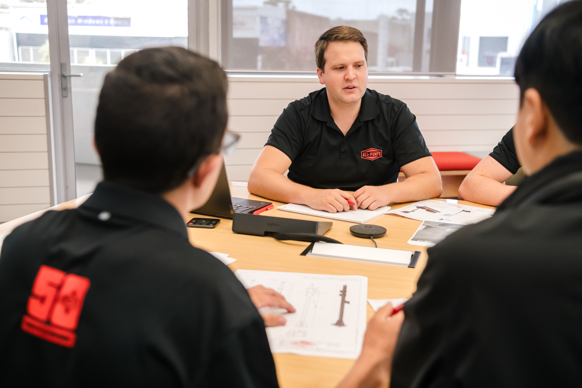 All-Pumps staff in a meeting room discussing engineering designs around a table, with sketches and laptops visible.