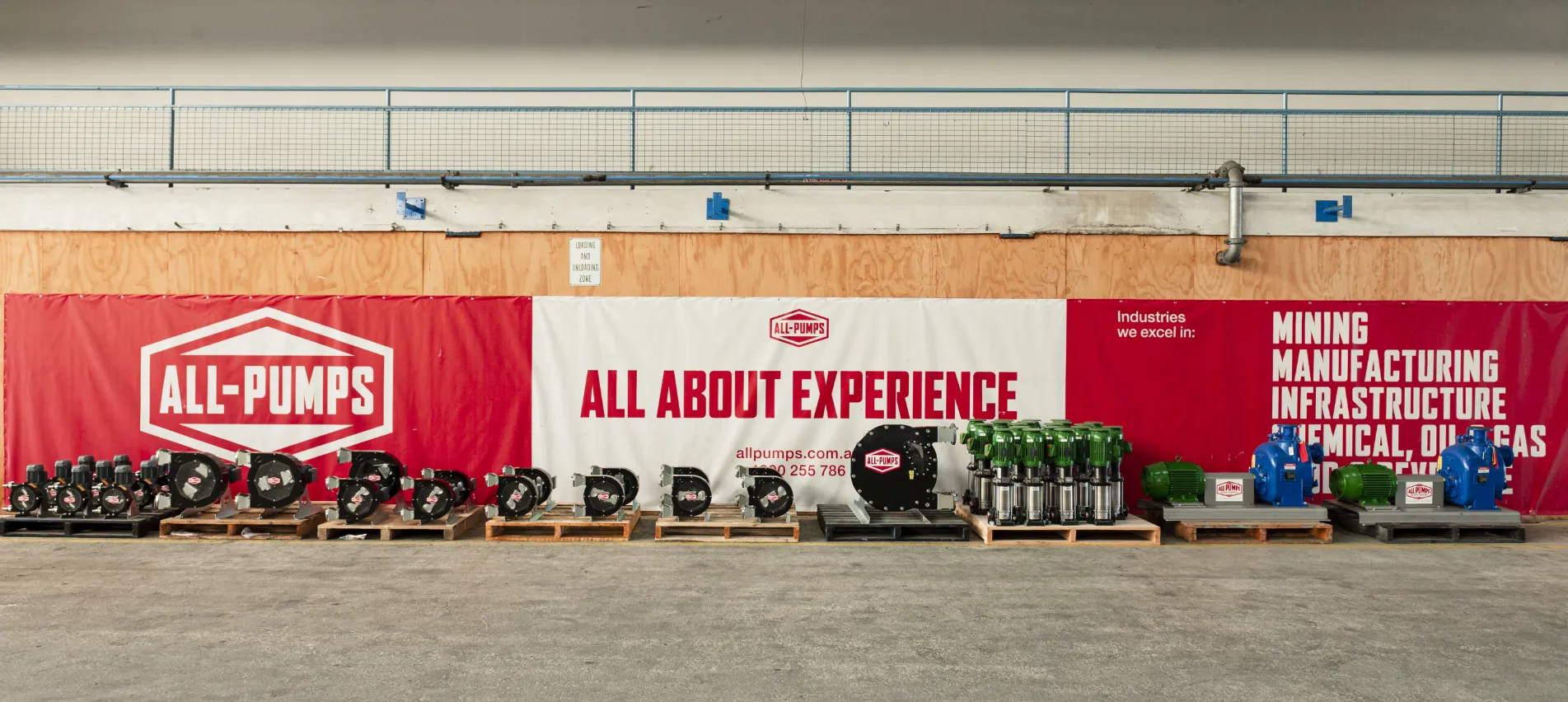 A lineup of industrial pumps displayed on pallets in front of a branded wall at All-Pumps, featuring the company’s slogan and the industries they serve, including mining and manufacturing.