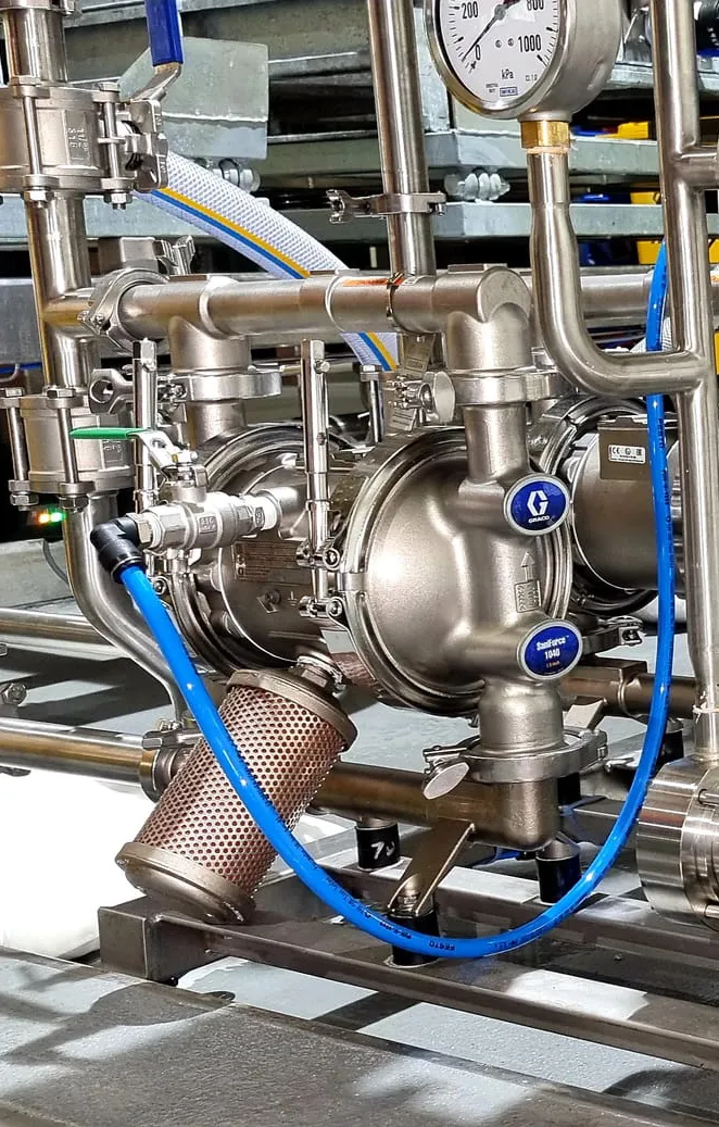 Stainless steel industrial pump system with pressure gauges, valves, and blue hoses in a clean processing facility.