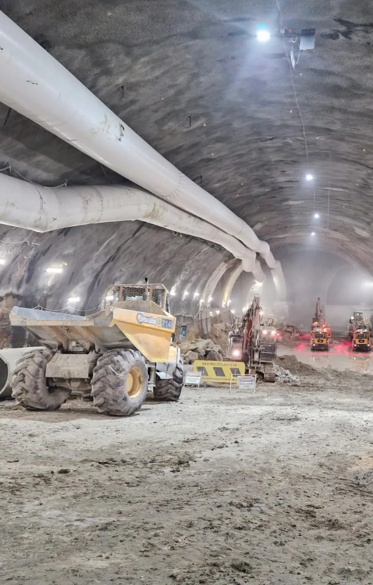 Inside a tunnel construction site, a large yellow dump truck and multiple excavators operate beneath overhead ventilation tubes.