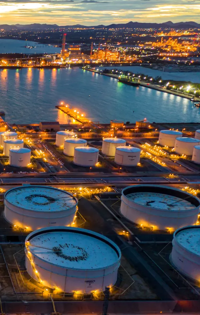 Aerial view of a large industrial port facility with illuminated storage tanks and refineries along a waterway at dusk.