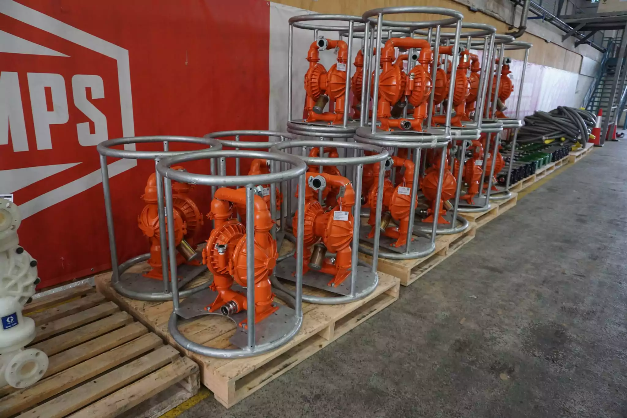 Multiple Wilden industrial pumps in protective metal frames, neatly arranged on wooden pallets inside a warehouse, with a red wall banner partially visible in the background.