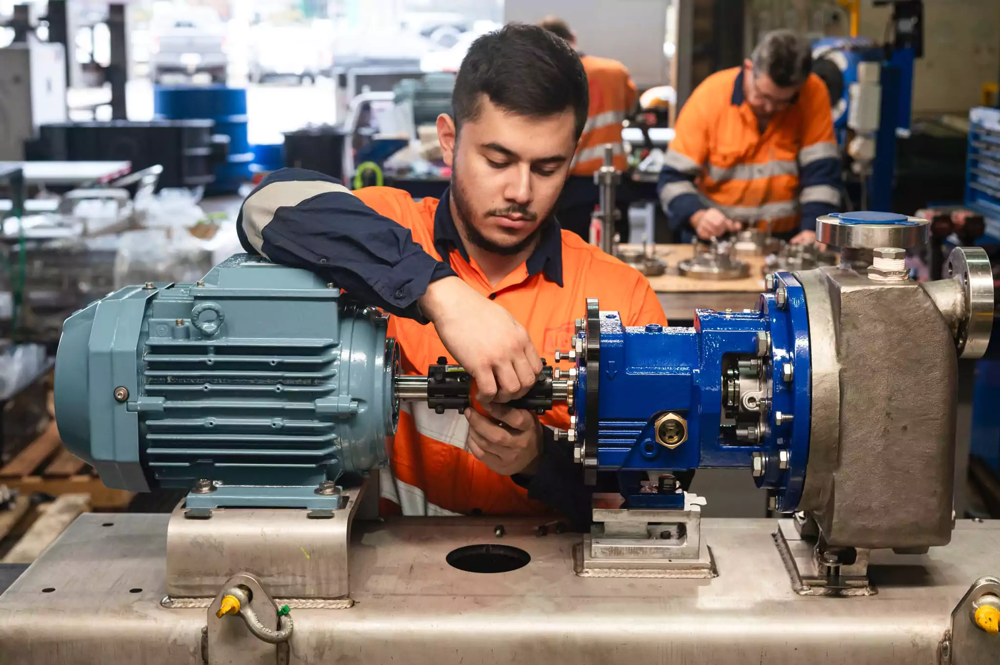 Technician assembling a Nexus industrial pump with a motor in a workshop, wearing high-visibility orange safety uniform, with other workers and equipment visible in the background.