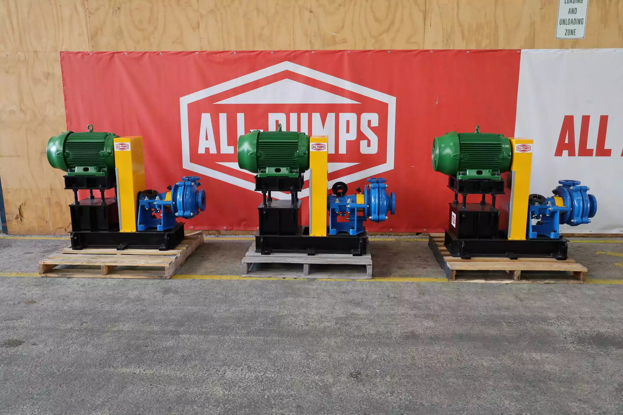 Three Nexus slurry pumps with green motors and blue pump casings, mounted on black base frames and wooden pallets, displayed in front of an All-Pumps banner inside a warehouse.